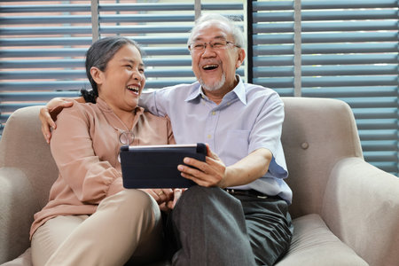 Happy smiling asian senior couple sitting on sofa and using tablet while online video call with friend or relative cousin at home living room. Internet information technology and lifestyle concept.の写真素材