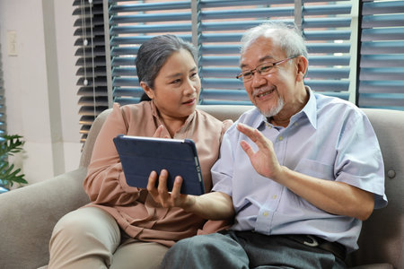 Happy smiling asian senior couple sitting on sofa and using tablet while online video call with friend or relative cousin at home living room. Internet information technology and lifestyle concept.の写真素材