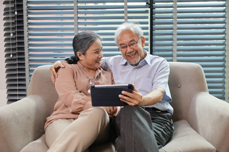 Happy smiling asian senior couple sitting on sofa and using tablet while online video call with friend or relative cousin at home living room. Internet information technology and lifestyle concept.の写真素材