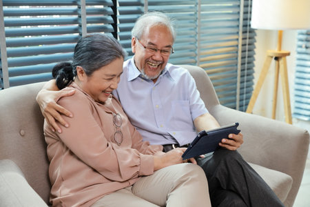 Happy smiling asian senior couple sitting on sofa and using tablet while online video call with friend or relative cousin at home living room. Internet information technology and lifestyle concept.の写真素材