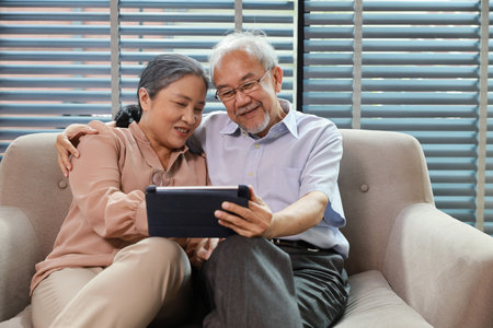 Happy smiling asian senior couple sitting on sofa and using tablet while online video call with friend or relative cousin at home living room. Internet information technology and lifestyle concept.の写真素材