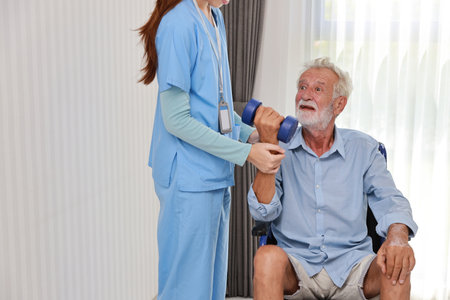 Happy caregiver helping senior elderly man lifting dumbbell and excercise with wheelchair at home or retirement house. Asian smiling nurse taking good help care and support of elder patient at house.の写真素材