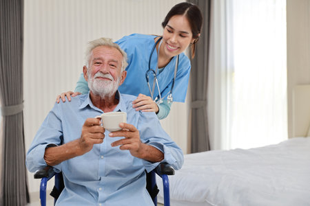 Happy caregiver giving senior caucasian man with wheelchair a cup of coffee or tea for breakfast in bedroom at retirement house. Asian smiling nurse taking good help care and support elder patient.の写真素材