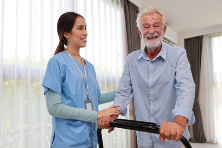 Happy caregiver helping senior elderly man walk with exercise machine in bedroom at home or retirement house. Asian smiling nurse taking good help care and support of elder patient at house.の写真素材