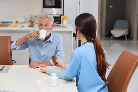 Happy caregiver sitting and helping senior caucasian man taking medicine after breakfast in living room at retirement house. Asian smiling nurse taking good help care and support elder patient at homeの写真素材