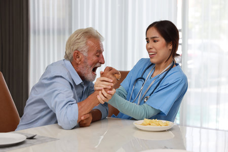 Happy caregiver try stopping senior caucasian man eating hamburger or junk food and taking medicine after breakfast in living room at retirement house. Unhealthy and junk food lifestyle conceptの写真素材