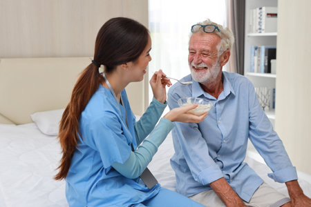 Happy caregiver sitting and feeding senior caucasian man on bed with rice porridge soup for breakfast at retirement house. Asian smiling nurse taking good help care and support elder patient at home.の写真素材