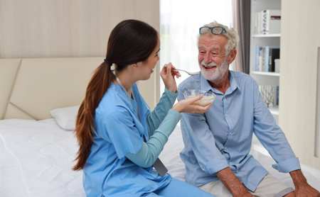 Happy caregiver feeding senior old man on bed with rice porridge soup for breakfast at retirement house. Asian smiling nurse taking good help care and support of elder patient at home.の写真素材