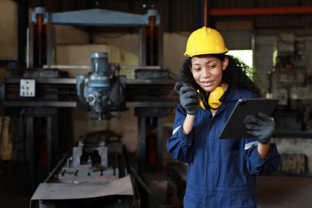 Technician engineer or worker woman in protective suit using walkie talkie radio with tablet while controlling or maintenance operation work lathe metal machine at heavy industry manufacturing factoryの写真素材