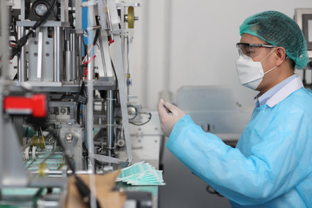 Worker in personal protective equipment or PPE inspecting quality of mask and medical face mask production line in factory, manufacturing industry and factory concept.の写真素材