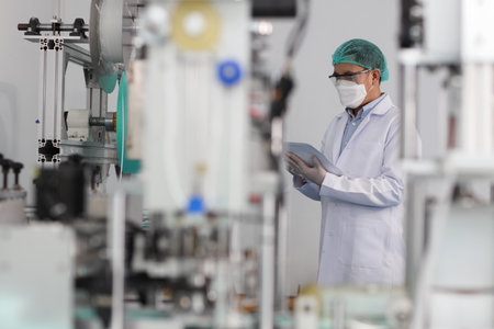 Worker man in personal protective equipment or PPE inspecting quality of mask and medical face mask production line in factory, manufacturing industry and factory concept.の写真素材