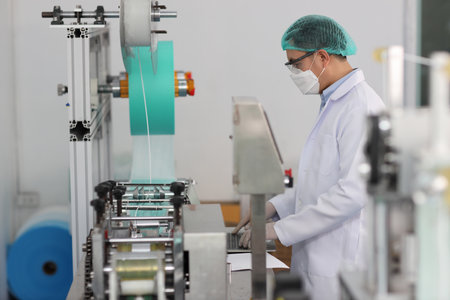 Worker man in personal protective equipment or PPE inspecting quality of mask and medical face mask production line in factory, manufacturing industry and factory concept.の写真素材