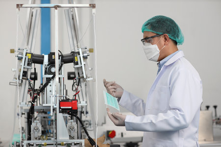 Worker man in personal protective equipment or PPE inspecting quality of mask and medical face mask production line in factory, manufacturing industry and factory concept.の写真素材