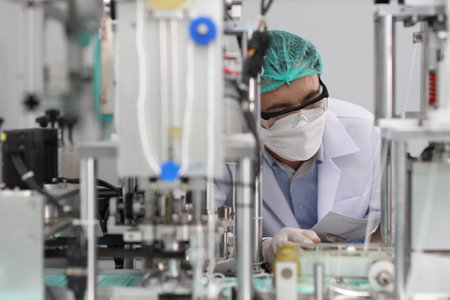 Worker in personal protective equipment or PPE inspecting quality of mask and medical face mask production line in factory, manufacturing industry and factory concept.の写真素材