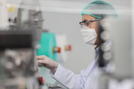 Worker man in personal protective equipment or PPE inspecting quality of mask and medical face mask production line in factory, manufacturing industry and factory concept.の写真素材