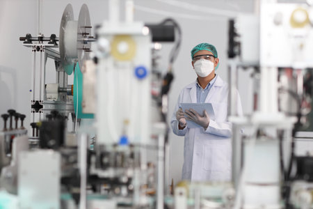 Worker in personal protective equipment or PPE inspecting quality of mask and medical face mask production line in factory, manufacturing industry and factory concept.の写真素材