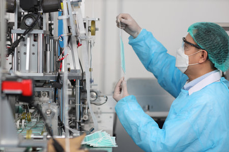 Worker in personal protective equipment or PPE inspecting quality of mask and medical face mask production line in factory, manufacturing industry and factory concept.の写真素材