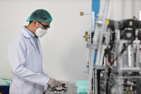 Worker in personal protective equipment or PPE inspecting quality of mask and medical face mask production line in factory, manufacturing industry and factory concept.の写真素材