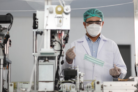 Worker in personal protective equipment or PPE inspecting quality of mask and medical face mask production line in factory, manufacturing industry and factory concept.の写真素材