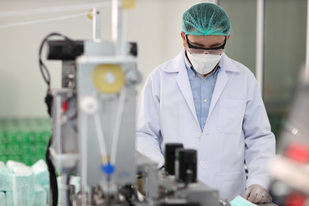 Worker in personal protective equipment or PPE inspecting quality of mask and medical face mask production line in factory, manufacturing industry and factory concept.の写真素材