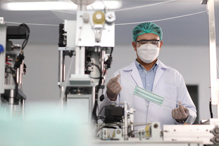 Worker man in personal protective equipment or PPE inspecting quality of mask and medical face mask production line in factory, manufacturing industry and factory concept.の写真素材