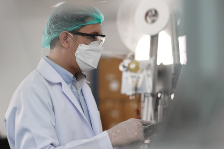 Worker in personal protective equipment or PPE inspecting quality of mask and medical face mask production line in factory, manufacturing industry and facの写真素材