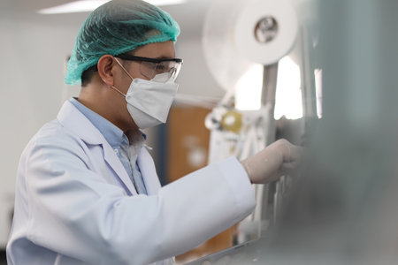 Worker in personal protective equipment or PPE inspecting quality of mask and medical face mask production line in factory, manufacturing industry and factory concept.の写真素材