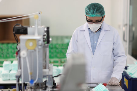 Worker man in personal protective equipment or PPE inspecting quality of mask and medical face mask production line in factory, manufacturing industry and factory concept.の写真素材
