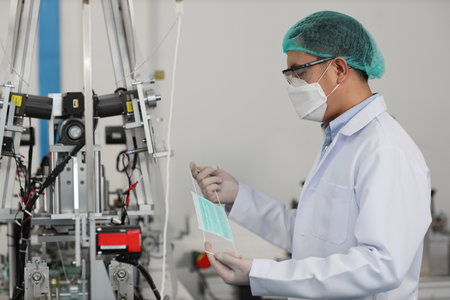 Worker man in personal protective equipment or PPE inspecting quality of mask and medical face mask production line in factory, manufacturing industry and factory concept.の写真素材