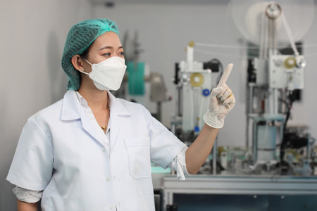 Worker in personal protective equipment or PPE inspection quality pointing something and looking away in mask and medical face mask production line factory, manufacturing industry and factory concept.の写真素材