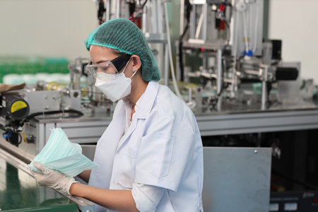 Worker woman in personal protective equipment or PPE inspecting quality of mask and medical face mask production line in factory, manufacturing industry and factory concept.の写真素材