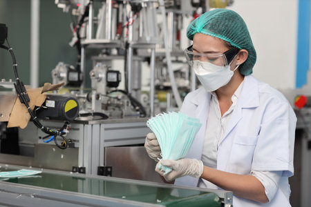 Worker woman in personal protective equipment or PPE inspecting quality of mask and medical face mask production line in factory, manufacturing industry and factory concept.の写真素材
