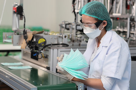 Worker woman in personal protective equipment or PPE inspecting quality of mask and medical face mask production line in factory, manufacturing industry and factory concept.の写真素材