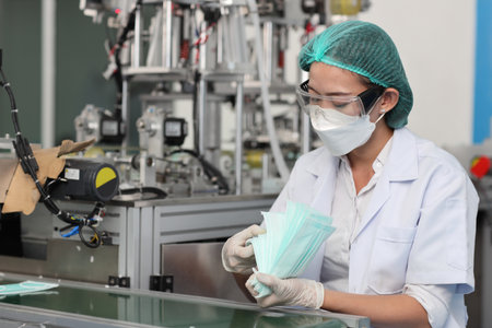 Worker woman in personal protective equipment or PPE inspecting quality of mask and medical face mask production line in factory, manufacturing industry and factory concept.の写真素材