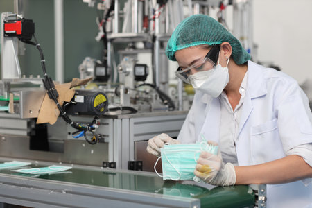 Worker woman in personal protective equipment or PPE inspecting quality of mask and medical face mask production line in factory, manufacturing industry and factory concept.の写真素材