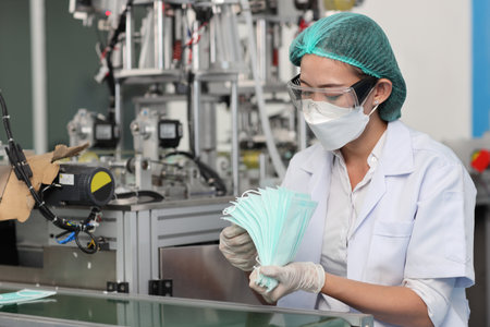 Worker woman in personal protective equipment or PPE inspecting quality of mask and medical face mask production line in factory, manufacturing industry and factory concept.の写真素材