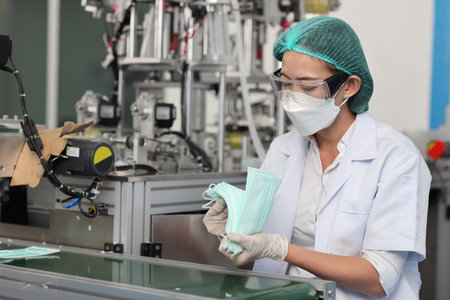 Worker woman in personal protective equipment or PPE inspecting quality of mask and medical face mask production line in factory, manufacturing industry and factory concept.の写真素材