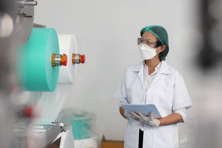 Worker woman in personal protective equipment or PPE inspecting quality of mask and medical face mask production line in factory, manufacturing industry and factory concept.の写真素材