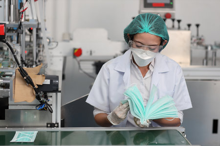 Worker woman in personal protective equipment or PPE inspecting quality of mask and medical face mask production line in factory, manufacturing industry and factory concept.の写真素材