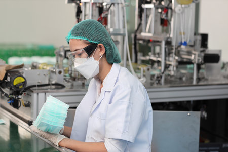 Worker woman in personal protective equipment or PPE inspecting quality of mask and medical face mask production line in factory, manufacturing industry and factory concept.の写真素材
