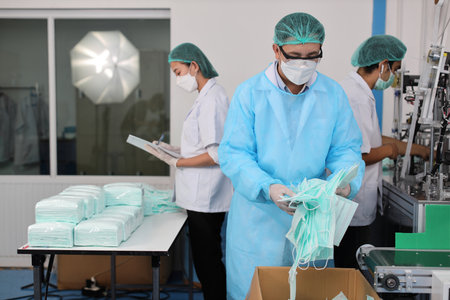 Worker man and woman in personal protective equipment or PPE inspecting checking quality of mask and medical face mask production line in factory, manufacturing industry and factory concept.の写真素材
