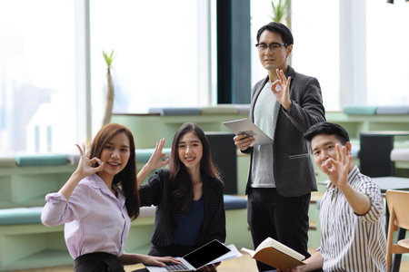 Collaborative process of young asian business people team working together discussing, researching, brainstorming and planning while showing OK symbol in business office building and city backgroundの写真素材