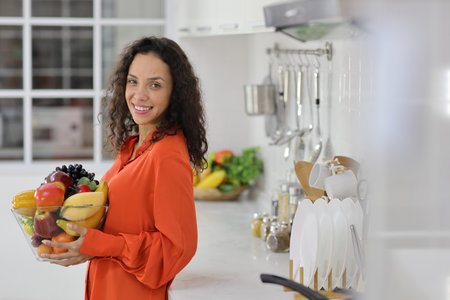 Happy latin woman standing and holding fruits basket while smiling and looking at camera in kitchen. Young female preparing delicious meal at home. Healthy and lifestyle conceptの写真素材