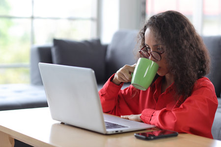 Smart and active latin woman sitting on sofa and using internet on computer with smart mobile phone while drinking coffee in living room. Lifestyle with technologyの写真素材