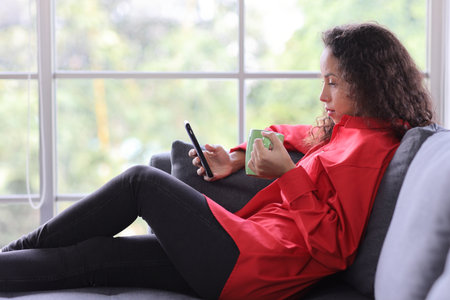 Happy smiling latin woman sitting on sofa while drinking coffee and using internet on smart mobile phone in living room. Lifestyle with technologyの写真素材