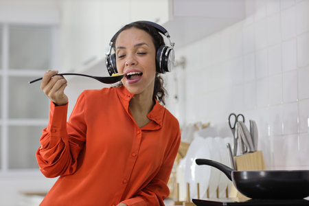 Happy latin woman cooking, smelling and tasting food in kitchen. Beautiful young female standing and preparing delicious meal with relaxation at home. Healthy and lifestyle conceptの写真素材