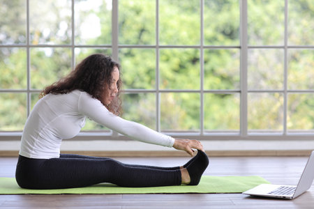 Sporty young adult woman sit on yoga mat practicing mediate yoga watching coach training video online from laptop. Healthy female doing exercise and taking lesson distant course among nature outdoorの写真素材
