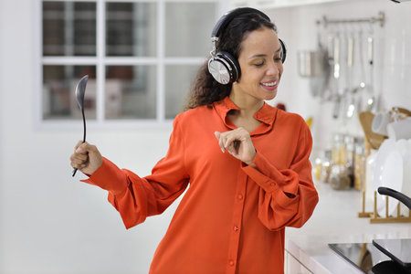 Happy latin woman cooking, smelling and tasting food while dancing in kitchen. Beautiful young female with headphone preparing delicious meal while listen music at home. Healthy and lifestyle conceptの写真素材