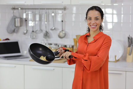 Happy latin woman cooking, smelling and tasting food in kitchen. Beautiful young female standing and preparing delicious meal with relaxation at home. Healthy and lifestyle conceptの写真素材