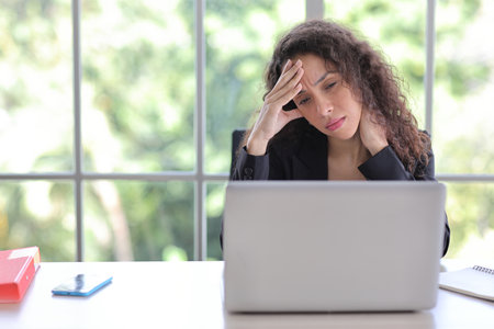 Adult businesswoman sitting and touching her head and looking at computer in office after bad news business failure or get fired and feeling discouraged, distraught and hopeless in modern officeの写真素材